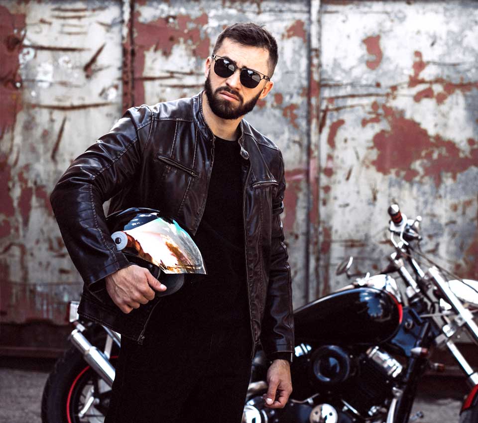 A man in a genuine black biker eather jacket and sunglasses stands next to a motorcycle, holding a helmet.