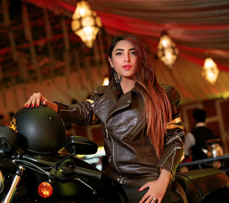 A woman with long brown hair sits on a black motorcycle. She wears a genuine brown leather jacket and red lipstick. The background is a red and gold event tent with hanging lights.